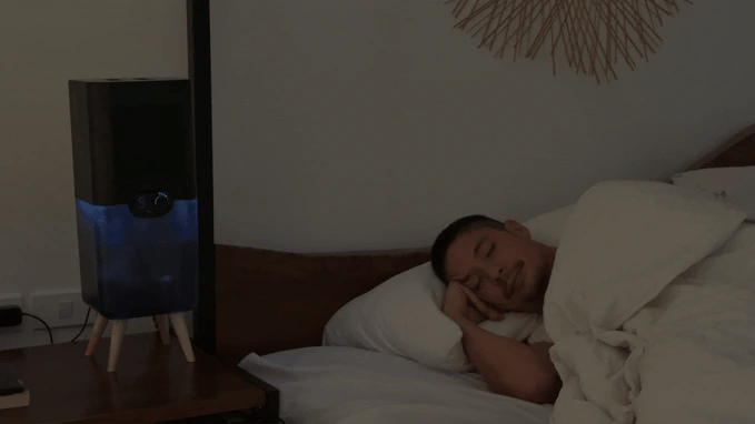 A bedroom at night, the lights dimming low while the Dreamora Luna unit glows softly on the nightstand beside a sleeping person